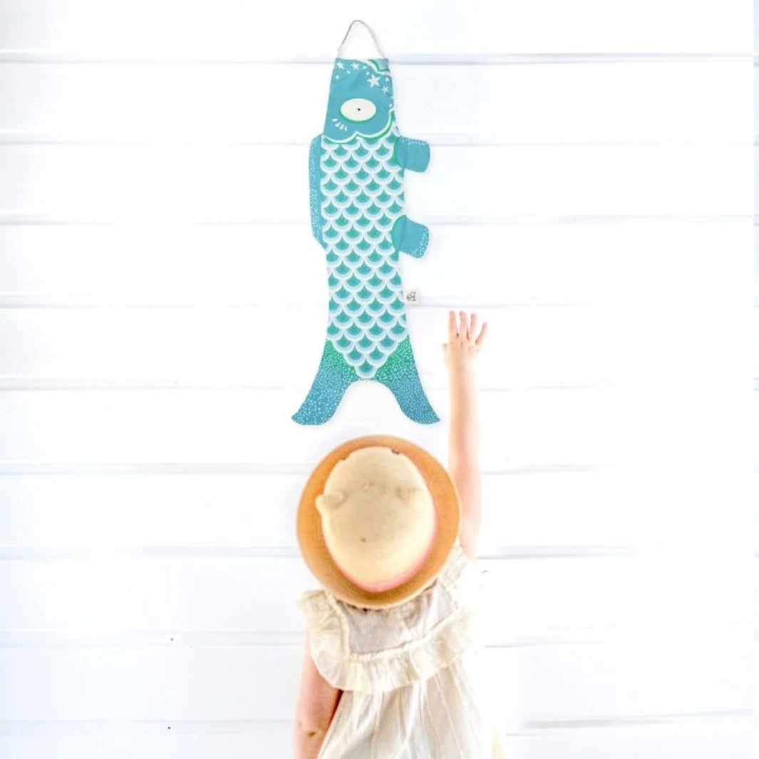 Kind mit Strohhut greift nach türkisem Koi-Windsack an heller Holzwand #farbe_Karibik
