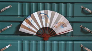 Aufgeklappter Vintage Mai-Ōgi mit drei gemalten Medaillons in Gold, Blau und Braun, rote Streben, auf petrolfarbener Vintage-Kommode